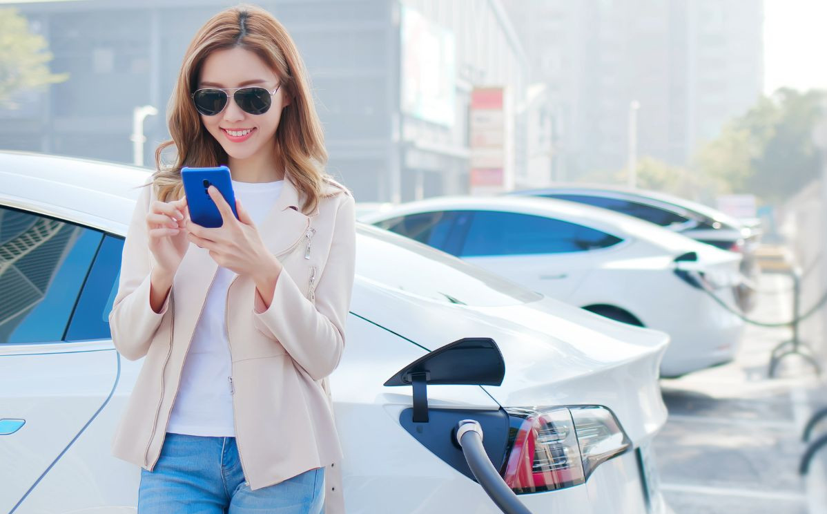 Une femme élégante tenant son téléphone portable à proximité d'une station de recharge, avec sa voiture électrique en arrière-plan