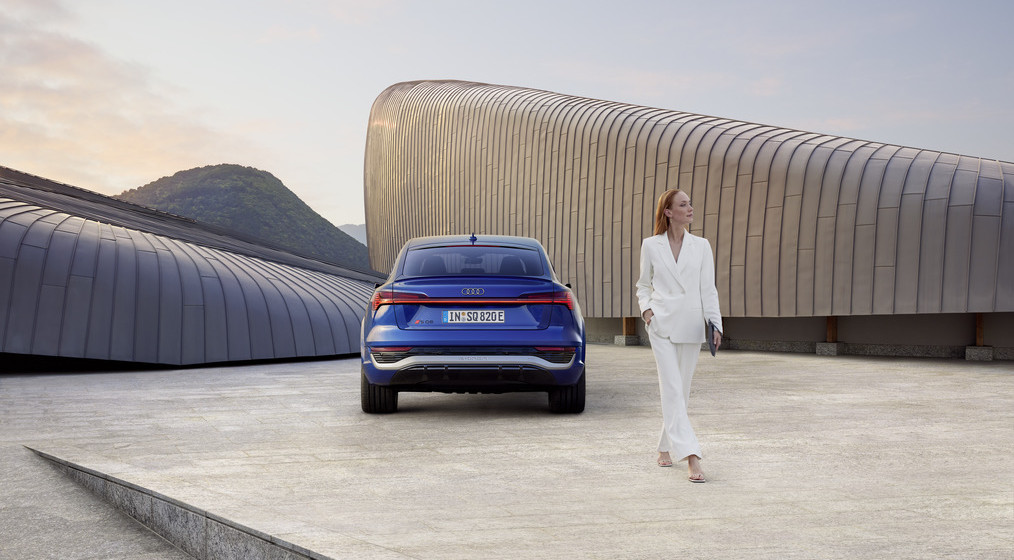 Arrière d'une voiture électrique Audi Q8 E-Tron bleue avec une dame habillée en blanc devant