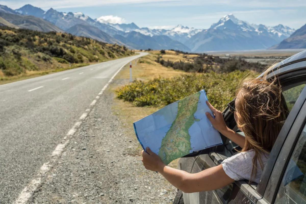 Femme sur le bas côté d'une route dans sa voiture électrique regardant une carte routière devant un paysage de montagne